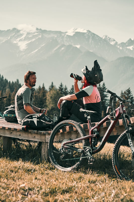 Two cyclists rest on a wooden platform. One drinks from a bottle, while the other sits casually. Their bicycles are parked nearby. Snow-capped mountains and a forest are in the background, under a clear sky.