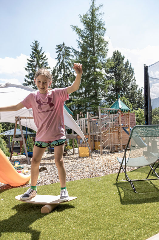 A child balances on a wooden board on a small cylinder in a backyard. They are wearing a pink shirt and shorts. The yard has green artificial grass, a slide, and a small climbing structure. Trees and mountains are visible in the background.