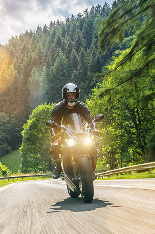A person wearing a helmet and black gear rides a motorcycle on a sunny road. The background features lush green trees and a forested hill, with sunlight streaming through the branches.