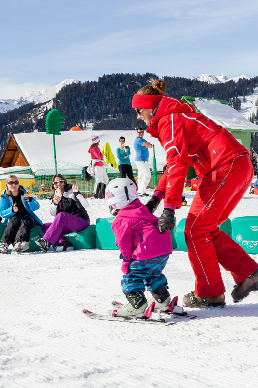 An instructor in a red suit helps a young child in a pink jacket ski down a snowy slope. Other people sit and watch against a backdrop of snow-covered mountains and clear blue sky.