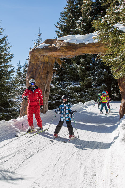 A group of skiers wearing winter gear, including helmets and goggles, ski down a snowy trail through a large wooden arch, surrounded by snow-covered trees on a sunny day.