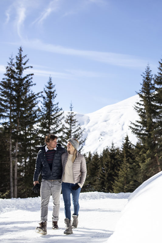A couple walks arm in arm on a snowy path surrounded by evergreen trees, with a snow-covered mountain in the background. They are both wearing winter jackets and boots, enjoying a clear, sunny day.