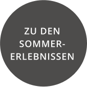 Ein dunkelgrauer Kreis mit weißem Text in deutscher Sprache mit der Aufschrift „ZU DEN SOMMER-ERLEBNISSEN“, der an den Charme von Alpenresorts erinnert.