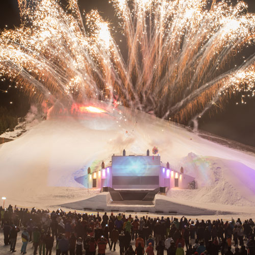 A large crowd watches vibrant fireworks illuminating the night sky over a snowy hill, marking the end of 3 fun ski days. The foreground features a stage with colorful lights, celebrating the festive atmosphere after a thrilling time on the slopes with a 2-day ski pass.