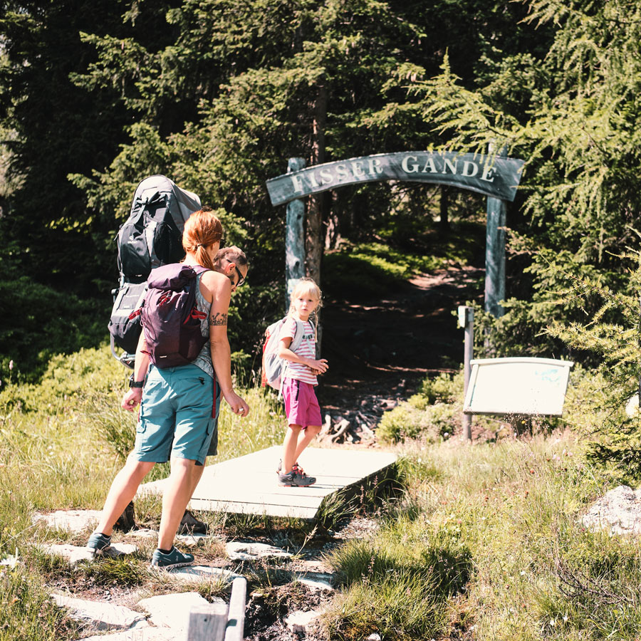 A family is hiking on a trail in a forested area. An adult and a child walk ahead, with another adult and child following, their backpacks packed for an adventure. As they approach the Risseu Gande entrance sign, they chat about staying 7 nights amidst the trees surrounding their path.
