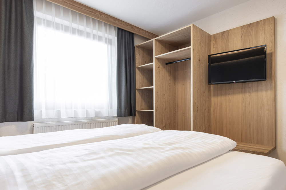 Ein ordentlich gemachtes Bett mit weißer Bettwäsche steht in einem Hotelzimmer im Neuen Apartment Gartenlodge in Ladis. Ein Holzschrank mit offenen Regalen wird von einem montierten Flachbildfernseher begleitet, während natürliches Licht durch ein mit weißen Vorhängen und dunklen Gardinen geschmücktes Fenster strömt.
