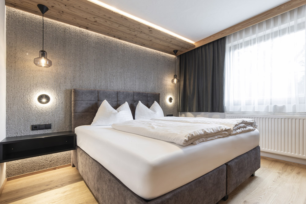 Ein modernes Schlafzimmer im Neuen Apartment Gartenlodge in Ladis verfügt über ein großes Bett mit weißen Kissen und Decken. Es besticht durch eine strukturierte graue Akzentwand, eine hölzerne Deckenplatte und hängende Pendelleuchten. Ein Fenster mit durchsichtigen und dunklen Vorhängen lässt natürliches Licht herein.