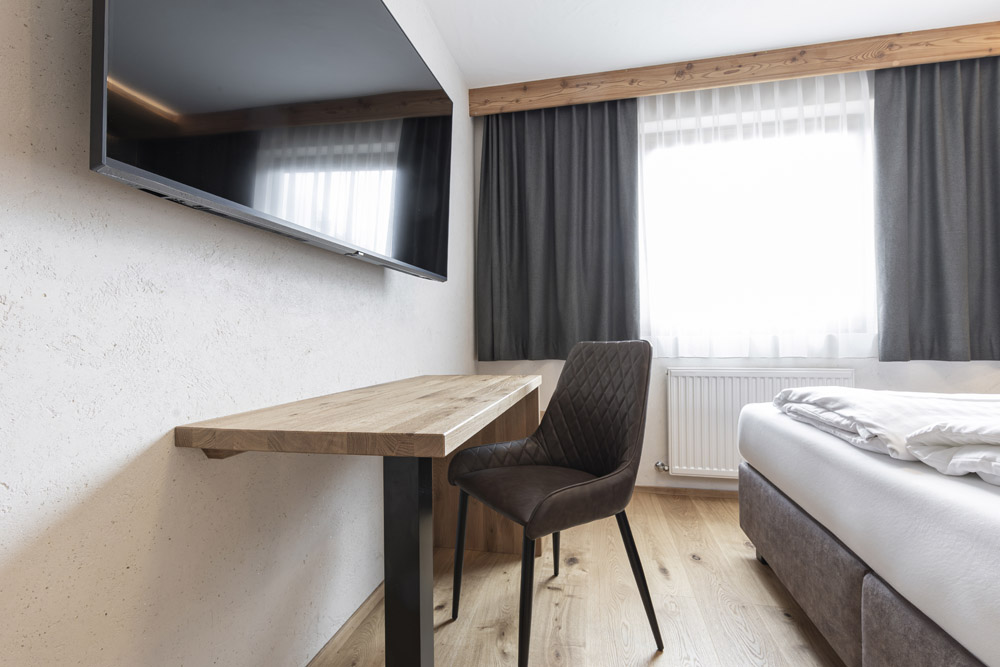 Modernes Hotelzimmer, das an eine Neue Apartment Gartenlodge in Ladis erinnert, mit einem Flachbildfernseher über einem Holzschreibtisch. Ein gepolsterter Stuhl steht vor dem Fernseher, während dunkle Vorhänge das Fenster einrahmen und Licht auf den hellen Holzboden und die schlichte Einrichtung fallen lassen.