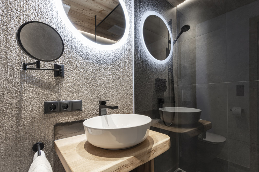Das moderne Badezimmer im Neuen Apartment Gartenlodge in Ladis verfügt über einen runden, von hinten beleuchteten Spiegel, einen runden Vergrößerungsspiegel und ein weißes Waschbecken auf einer Holzplatte. Strukturierte Betonwände und ein Duschbereich in Kombination mit schwarzen Armaturen sorgen für einen eleganten Kontrast.