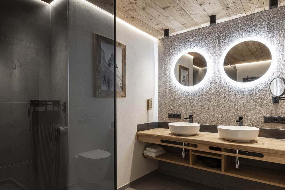 Dieses moderne Badezimmer im Neuen Apartment Gartenlodge in Ladis verfügt über strukturierte Wände und eine Holzdecke. Über einem Doppelwaschtisch befinden sich zwei runde Spiegel mit Hintergrundbeleuchtung, während links eine Glasduschkabine zu sehen ist. Handtücher liegen ordentlich darunter und Kunstwerke schmücken die Wand.