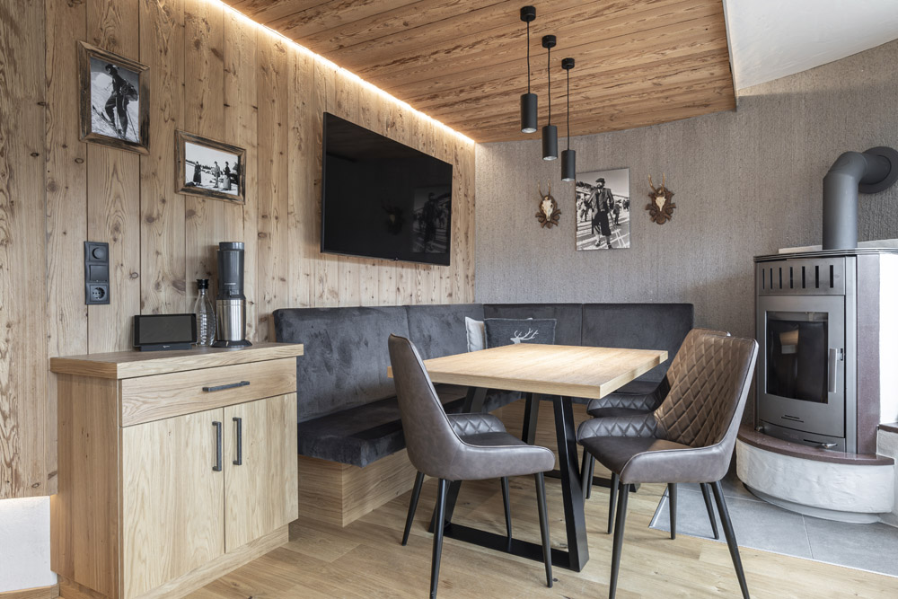 Eine gemütliche Essecke im Neuen Apartment Gartenlodge in Ladis verfügt über Holzwände und -decke, eine Eckbank und Lederstühle um einen hellen Holztisch. Ein Flachbildfernseher und Vintage-Fotos schmücken die Wand, während in der Ecke ein moderner Ofen für Wärme sorgt.