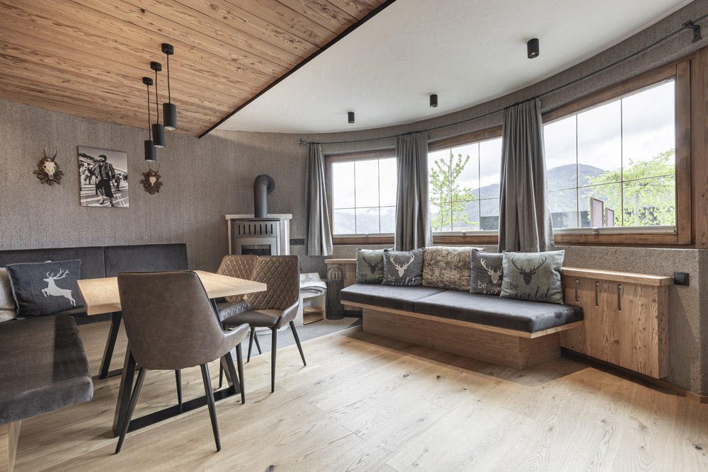 Ein gemütlicher, rustikaler Ess- und Wohnbereich im Neuen Apartment Gartenlodge in Ladis verfügt über Holzböden und -decken. Ein Tisch mit Stühlen, ein Holzofen, eine gepolsterte Bank mit dekorativen Kissen, große Fenster mit Bergblick und Kunstwerke mit Naturmotiven schmücken die Wände.