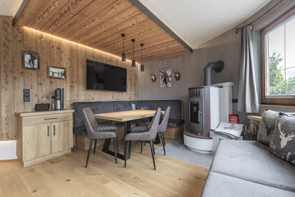 Gemütliche Holzhütteneinrichtung, die an eine Neue Apartment Gartenlodge in Ladis erinnert, mit einem kleinen Esstisch mit Stühlen, einem Ecksofa mit Kissen mit Hirschmotiv, einem an der Wand montierten Fernseher, einem modernen Ofen, einer Holzdecke und einem großen Fenster. Genießen Sie das warme, rustikale Ambiente und das natürliche Licht.