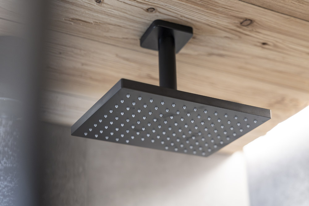 Ein moderner quadratischer schwarzer Duschkopf ist an einer Holzdecke im Neuen Apartment Gartenlodge in Ladis montiert. Er verfügt über mehrere kleine Wasserdüsen und ergänzt das minimalistische Badezimmerdesign mit grauen Wänden.