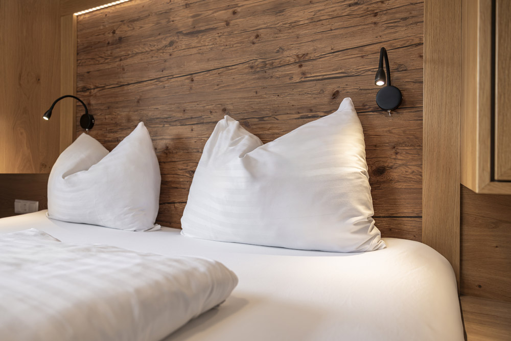Ein gemütliches Bett mit weißen Kissen und einer Bettdecke steht vor einem rustikalen Holzkopfteil, perfekt für Ihren Aufenthalt in einer Lodge in Serfaus-Fiss-Ladis für 2-4 Personen. Auf jeder Seite sind zwei schwarze, verstellbare Leselampen angebracht, die die warme und einladende Atmosphäre des Zimmers verstärken.