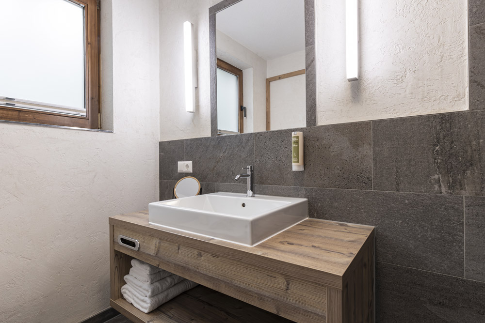 Dieses minimalistische Badezimmer für 2-4 Personen erinnert an eine Hütte in Serfaus-Fiss-Ladis und verfügt über einen hölzernen Waschtisch mit einem weißen, rechteckigen Waschbecken und einem modernen Wasserhahn. Darüber hängt ein großer Spiegel mit schmalen vertikalen Lichtern. Handtücher sind ordentlich auf dem offenen Regal darunter gestapelt und ergänzen das ruhige Ambiente.