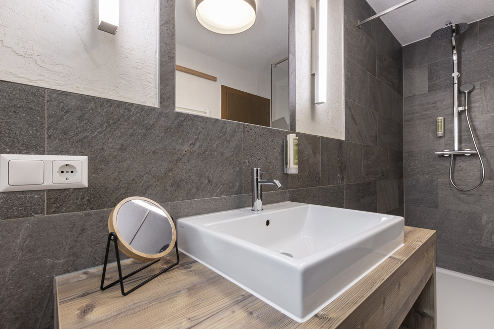 Diese moderne Lodge in Serfaus-Fiss-Ladis für 2-4 Personen verfügt über ein elegantes Badezimmer mit einem quadratischen weißen Waschbecken auf einer Holzplatte, ergänzt durch einen an der Wand montierten Wasserhahn und einen runden Vergrößerungsspiegel. Grau geflieste Wände, Deckenbeleuchtung und eine ebenerdige Dusche vervollständigen den eleganten Raum.