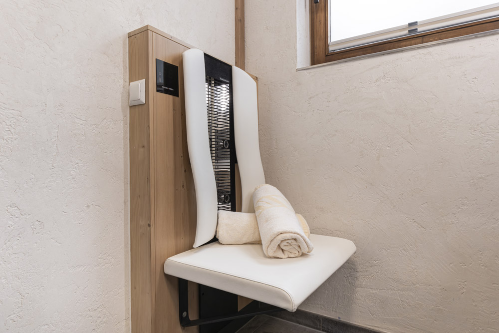 Ein moderner, weißer und hölzerner Infrarot-Heizstuhl mit schlichtem Design steht in einer hellen Ecke am Fenster. Aufgerollte beige Handtücher liegen ordentlich auf dem Sitz und schaffen eine Spa-ähnliche Oase, die an eine ruhige Lodge in Serfaus-Fiss-Ladis für 2-4 Personen erinnert.