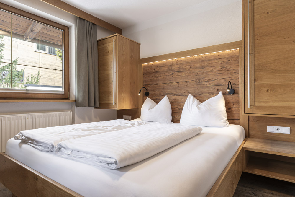 Eine gemütliche Lodge in Serfaus-Fiss-Ladis für 2-4 Personen verfügt über ein Schlafzimmer mit einem Doppelbett aus Holz, weißer Bettwäsche und zwei Kissensets. Das passende Kopfteil und die Schränke aus Holz verstärken den Charme, während ein Fenster mit Vorhängen natürliches Licht hereinlässt. Ein kleines Nachttischregal und Leselampen runden den Raum ab.