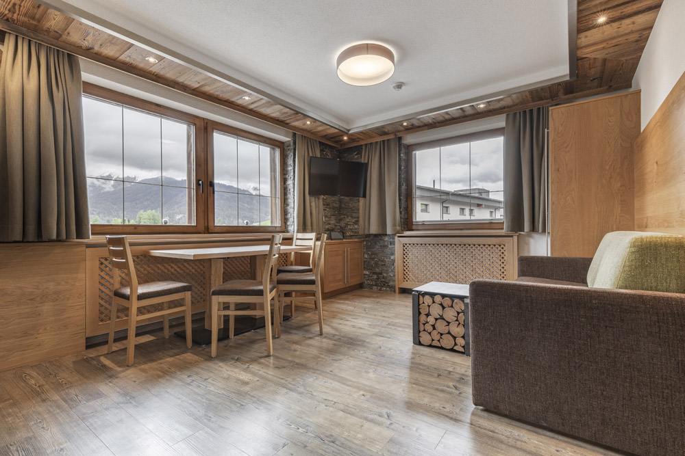 Modernes Wohnzimmer mit großen Fenstern und Bergblick, das an eine gemütliche Hütte in Serfaus-Fiss-Ladis für 2-4 Personen erinnert. Es verfügt über einen hölzernen Esstisch mit vier Stühlen, ein braunes Sofa mit Polstern und einen kleinen Tisch mit gestapelten Holzscheiten. Die Holzvertäfelung umrahmt den an der Wand montierten Fernseher.