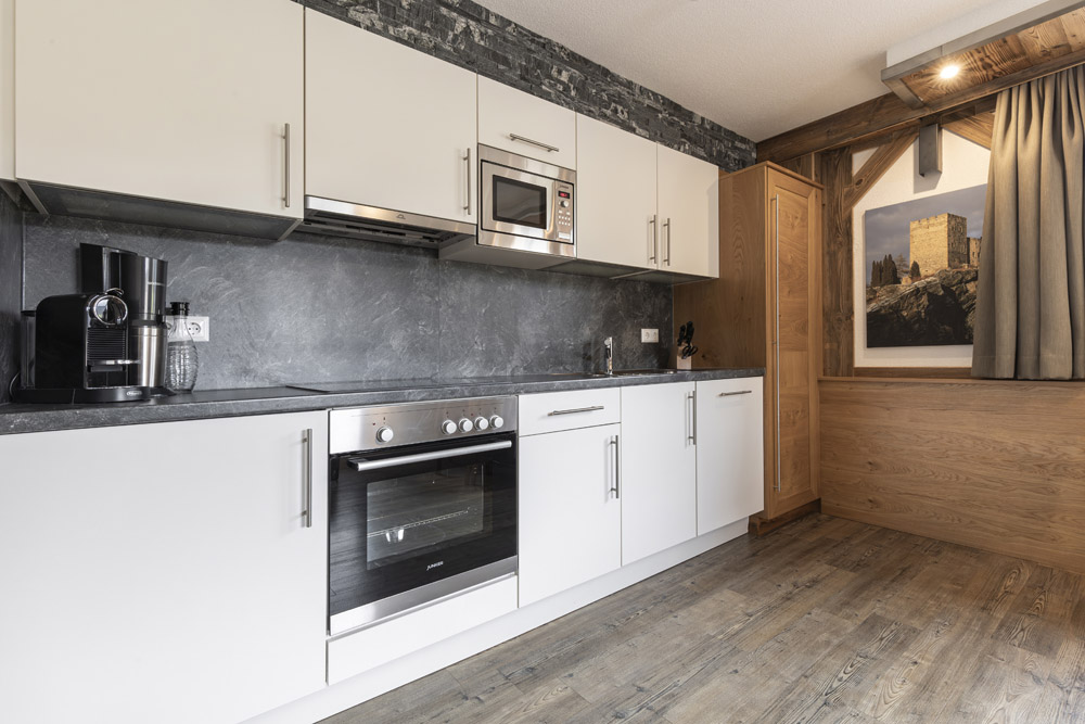 Moderne Küche mit weißen Schränken, Edelstahlofen und Mikrowelle. Auf der Arbeitsplatte stehen eine Kaffeemaschine und ein Glasgefäß. Dunkle Steinrückwand und Holzpaneelakzente sorgen für rustikalen Charme. Diese gemütliche Lodge in Serfaus-Fiss-Ladis für 2-4 Personen verfügt über ein Fenster mit atemberaubendem Blick auf die Burg.