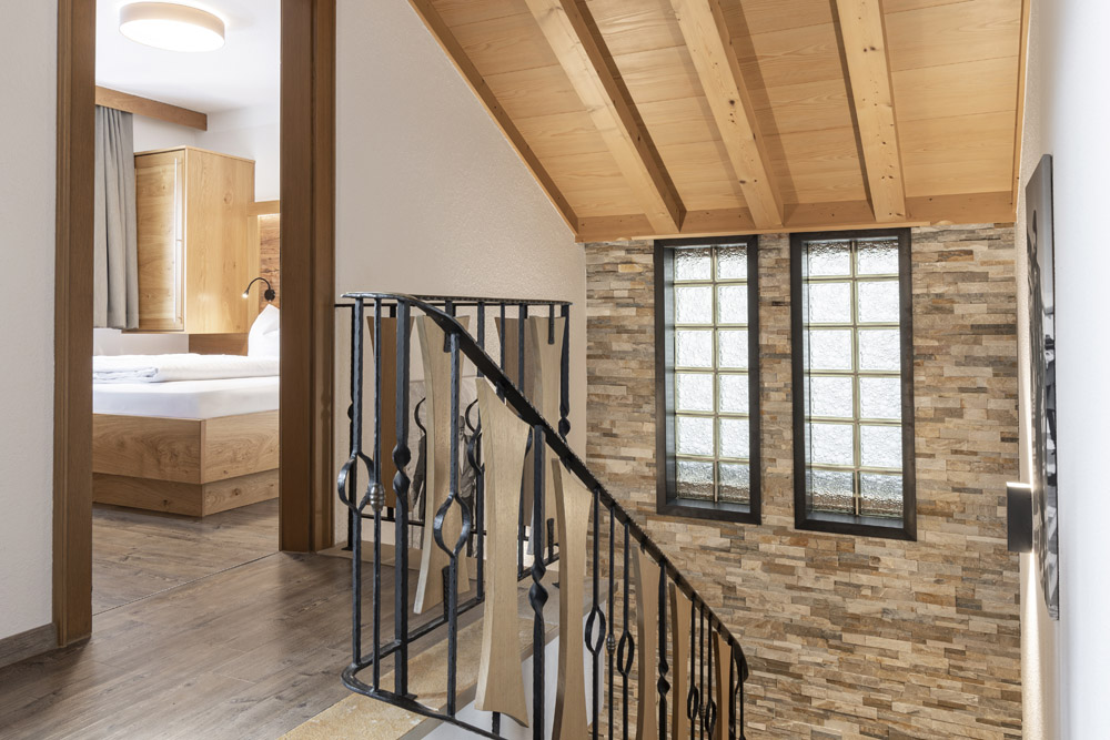 Eine gemütliche Lodge in Serfaus-Fiss-Ladis für 2-4 Personen verfügt über eine Holztreppe mit schwarzem Metallgeländer, die zu einem Zimmer mit einem Bett führt. Der Raum hat eine Akzentwand aus Stein und große Fenster, während eine Holzdecke und ein Holzboden den rustikalen Look vervollständigen.