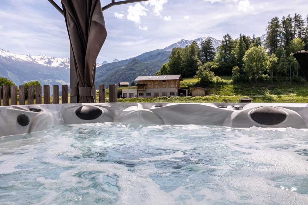 Ein sprudelnder Whirlpool mit einer Bergkulisse im Hintergrund rundet diese ruhige Szene ab. Schneebedeckte Gipfel erheben sich über dem Family Chalet für 4-6 Personen in Ladis, wo grüne Bäume und ein rustikaler Zaun ein gemütliches Holzhaus unter einem teilweise bewölkten Himmel umrahmen.