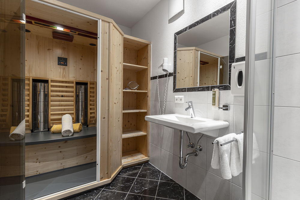Ein modernes Badezimmer im Alpenapart, ideal für 4-6 Personen, verfügt über eine Holzsauna, ein weißes Waschbecken und einen Wandspiegel. Der schwarz geflieste Boden bildet einen Kontrast zu den hellgrauen Wänden. Handtücher sind in der Sauna ordentlich gefaltet, eines hängt bequem neben dem Waschbecken.