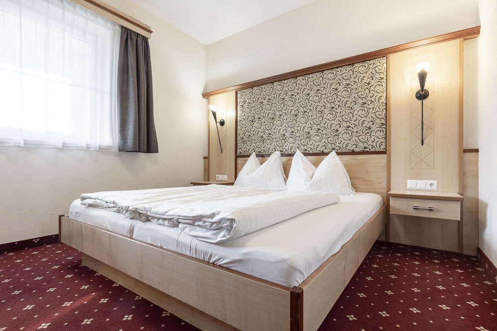 Im Alpenapart Apartment für 4-6 Personen steht ein ordentlich gemachtes Doppelbett mit weißer Bettwäsche und dreieckigen Kissen vor einem verzierten Kopfteil. Sanftes Licht von Wandleuchten und ein Fenster mit transparenten Vorhängen erhellen den Raum. Der rote Teppich ist mit kleinen weißen Mustern versehen.