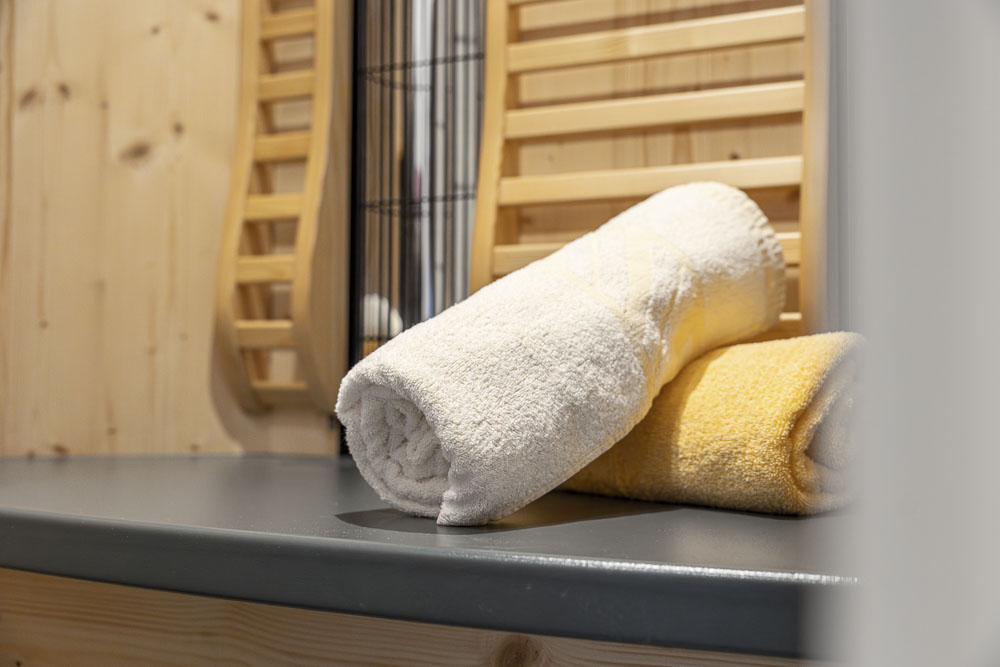 Zwei aufgerollte Handtücher, ein weißes und ein gelbes, liegen auf einer grauen Fläche mit Holzwandpaneelen und einem vertikalen Metallgestell im Hintergrund und fangen das gemütliche Badezimmerambiente eines Alpenapart-Apartments für 4–6 Personen ein.