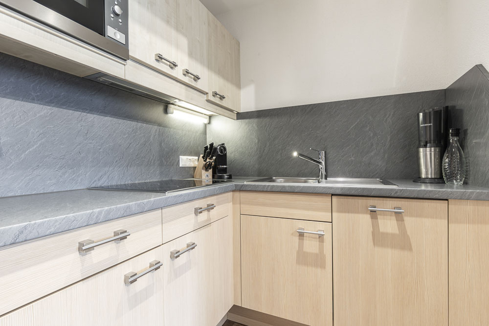 Diese moderne Küche in einem Apartment für 4-6 Personen im Alpenapart in Ladis verfügt über helle Holzschränke und graue Steinarbeitsplatten mit passender Spritzwand. Sie umfasst ein eingebautes Kochfeld, eine Edelstahlspüle mit Wasserhahn, ein Messerset und Geräte wie einen Mixer. Minimalistische Beleuchtung sorgt für Eleganz unter den Oberschränken.