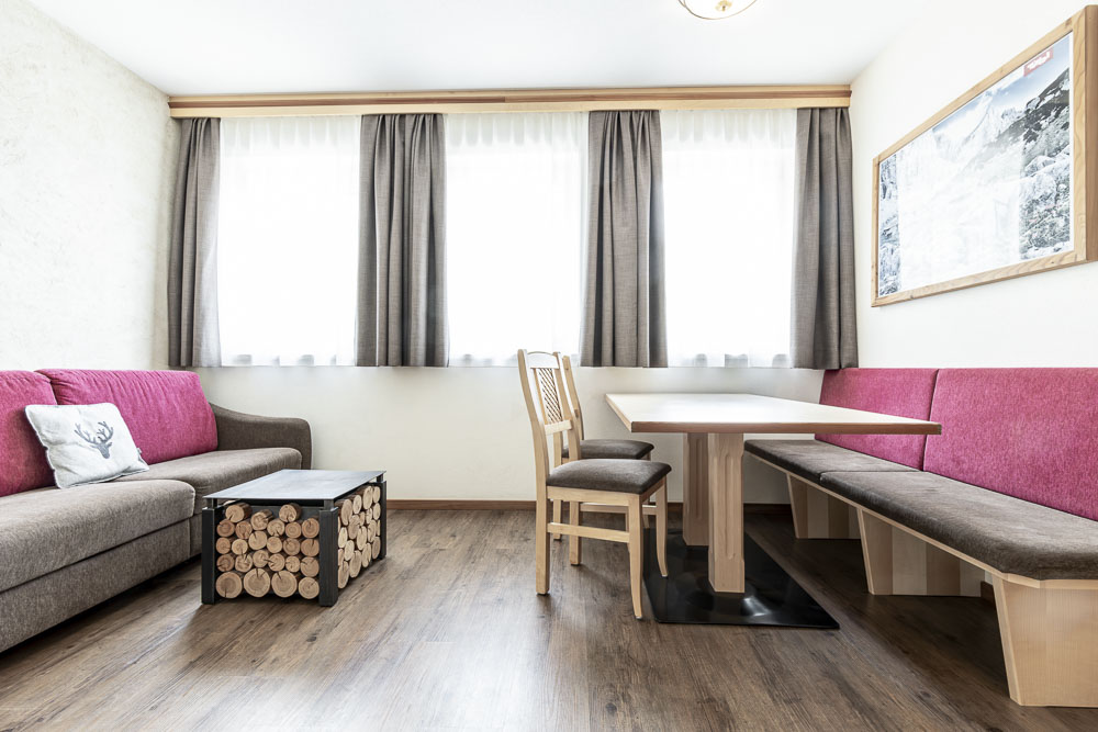 Modernes Wohnzimmer in einem Apartment für 4 Personen im Alpenapart in Ladis, mit hellem Holzboden, einem braunen und rosa Sofa und einem hölzernen Couchtisch mit gestapeltem Holzscheitdesign. Genießen Sie das Essen am Tisch mit Stühlen an großen Fenstern, die mit transparenten und grauen Vorhängen bedeckt sind, und einem gerahmten Bild, das die Wand akzentuiert.