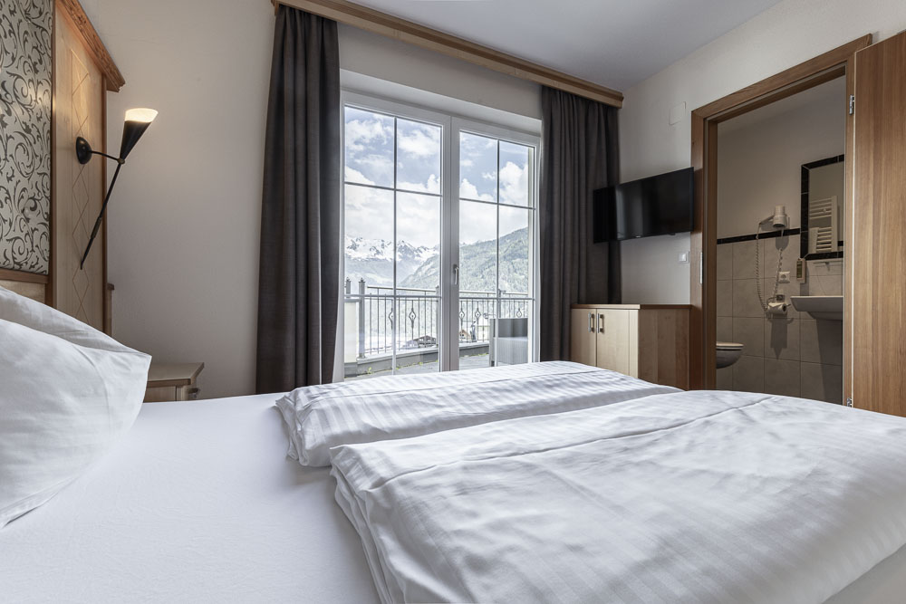 Das helle Apartment für 4 Personen im Alpenapart in Ladis verfügt über ein ordentlich gemachtes Bett mit Blick auf große Fenster. Genießen Sie den Blick auf schneebedeckte Berge, einen an der Wand montierten Fernseher, Holzmöbel und eine offene Tür, die zum Badezimmer führt.