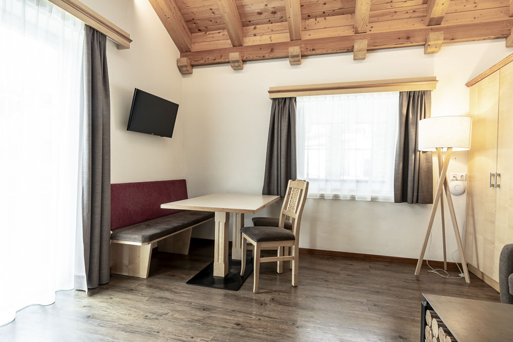 Ein gemütliches Apartmentzimmer mit Holzbalken an der Decke verfügt über einen an der Wand montierten Fernseher, einen Holztisch mit einer roten gepolsterten Bank und einem Stuhl, eine hohe Stehlampe und ein großes Fenster mit Vorhängen. Der Raum verfügt über Holzböden und ist in natürliches Licht getaucht.