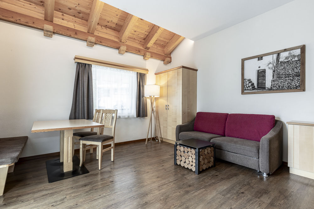 In einem gemütlichen Wohnzimmer mit Holzdecke und -boden steht ein kastanienbraunes Sofa neben einem kleinen Tisch, auf dem Holzscheite gestapelt sind. Der Raum umfasst einen Esstisch mit zwei Stühlen am Fenster und eine hohe Lampe steht neben dem Schrank. An der Wand hängt ein gerahmtes Foto.