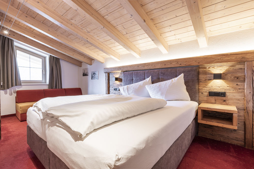 Ein gemütliches Schlafzimmer in einem Apartment für 4-6 Personen in Ladis, Tirol, verfügt über ein großes Bett unter einer schrägen Holzdecke. Weiße Bettwäsche bildet einen Kontrast zum rustikalen Holzkopfteil. Ein roter Teppich bedeckt den Boden und ein Fenster mit Vorhängen lässt von links natürliches Licht herein.