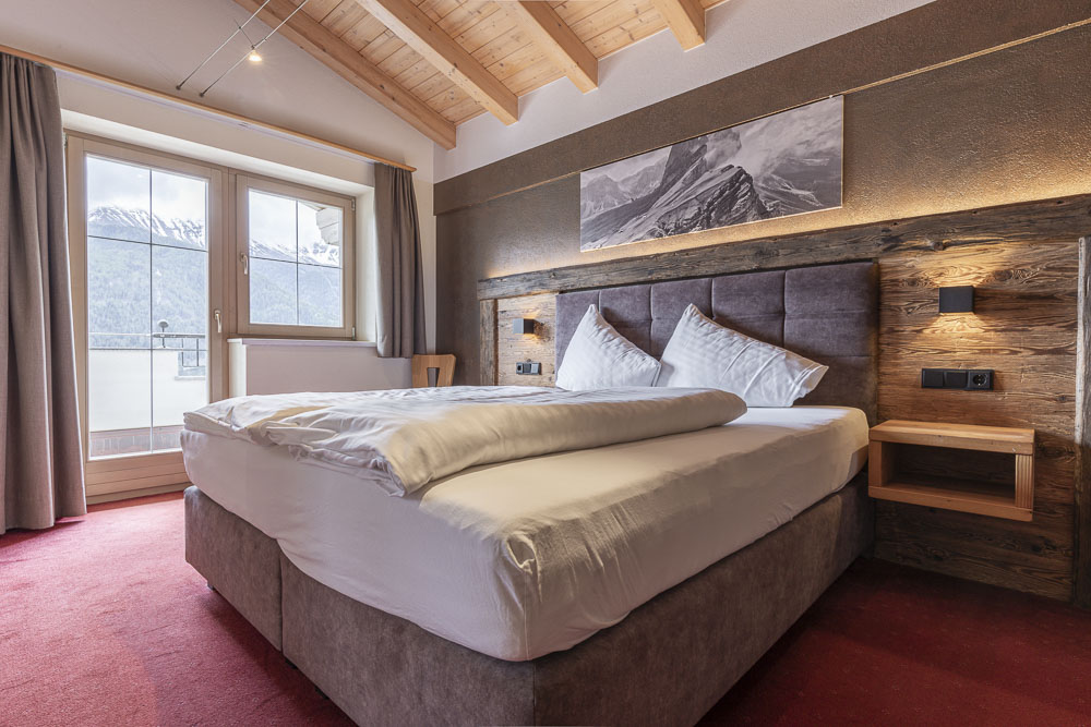 Gemütliches Schlafzimmer in einem Apartment für 4-6 Personen in Ladis, Tirol, mit einem großen Bett mit weißer Bettwäsche und einem rustikalen Kopfteil aus Holz. Ein Kunstwerk mit Bergmotiven schmückt die schräge Deckenwand. Das Fenster bietet einen atemberaubenden Blick auf schneebedeckte Berge, und sanftes Licht unterstreicht die Wärme des Zimmers.