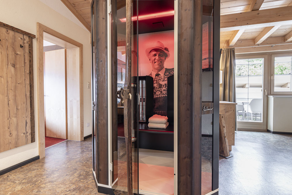 Ein gemütliches, rustikales Zimmer in einem Apartment für 4-6 Personen in Ladis Tirol verfügt über eine hölzerne Infrarotsaunakabine. Die offene Tür gibt den Blick auf einen Sitzbereich und ein Schwarzweißfoto eines lächelnden älteren Mannes frei, während natürliches Licht durch das Fenster strömt und die Holzbalken und den Holzboden hervorhebt.