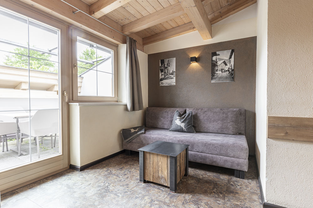 Ein gemütliches Apartment für 4-6 Personen in Ladis, Tirol, verfügt über einen Raum mit Holzdecke und Fliesenboden. Ein graues Sofa mit Kissen ergänzt den kleinen Couchtisch aus Holz. Zwei gerahmte Fotos schmücken die braune Akzentwand und eine Glastür öffnet sich zu einem Sitzbereich im Freien.