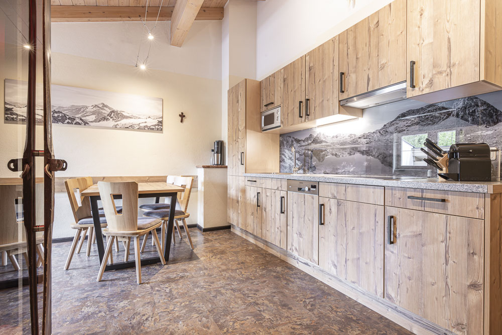 Eine gemütliche Küche in einem Apartment für 4-6 Personen in Ladis Tirol, mit Holzschränken und einer Küchenrückwand mit Bergmotiven. Links ergänzt ein Essbereich mit Holztisch und Stühlen das warme, rustikale Ambiente mit natürlichem Licht und freiliegenden Deckenbalken.