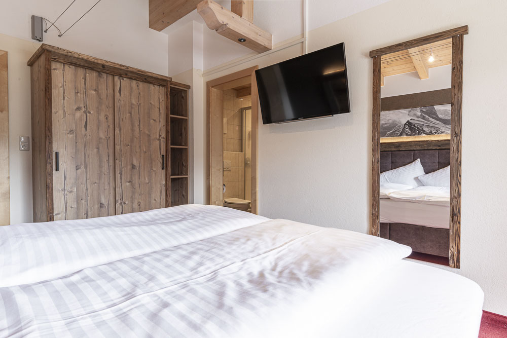Dieses gemütliche Schlafzimmer in einem Apartment für 4-6 Personen in Ladis Tirol verfügt über ein großes Bett, Holzmöbel und einen an der Wand montierten Fernseher. Ein großer Spiegel reflektiert das Bergmotiv, verstärkt das rustikale Ambiente mit Holzakzenten und bietet einen teilweisen Blick ins Badezimmer.