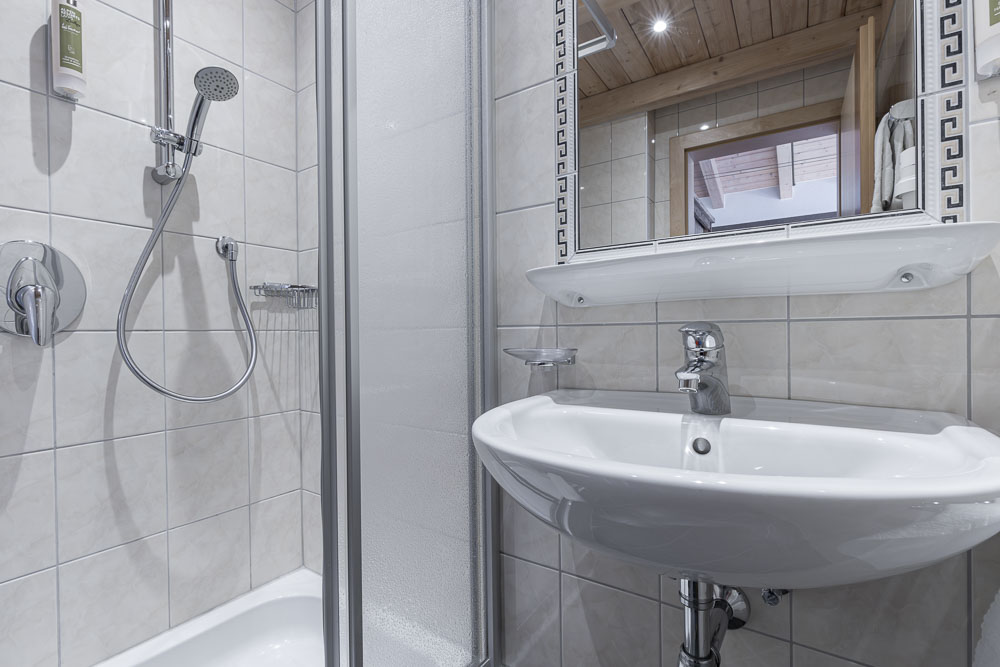 Ein kleines Badezimmer im Apartment für 4-6 Personen in Ladis Tirol verfügt über ein weißes Waschbecken mit einem Chromhahn unter einem Spiegel. Links davon befindet sich eine Duschkabine mit Glastür und Handbrause, während beige geflieste Wände und eine Holzdecke das gemütliche Design vervollständigen.