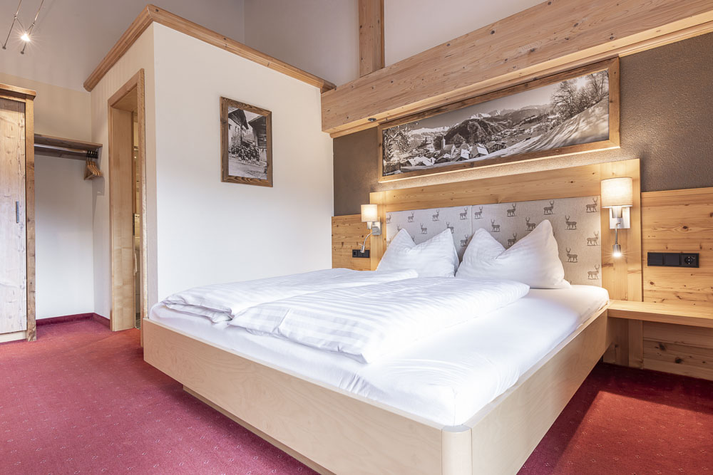 Gemütliches Hotelzimmer im Stil einer Wohnung für 4-5 Personen in Ladis, mit großem Bett mit weißer Bettwäsche, Kopfteil und Paneelen aus Holz. Ein roter Teppich bedeckt den Boden, während ein gerahmtes Landschaftsfoto die rustikale Einrichtung unterstreicht. Schrank und Türöffnung sind links zu sehen.