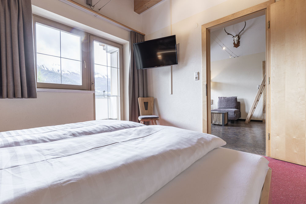 Das Apartment 6 für 4-5 Personen in Ladis ist ein gemütlicher Rückzugsort und bietet ein einladendes Hotelzimmer mit einem Bett mit karierter Bettdecke und malerischem Bergblick. Ein Flachbildfernseher schmückt die Wand, während eine offene Tür Sie in einen charmanten Sitzbereich mit Geweihdekor einlädt.