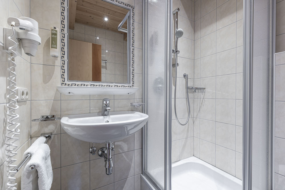 Dieses moderne Badezimmer im Apartment 6 für 4-5 Personen in Ladis verfügt über beigefarbene Fliesen, ein wandmontiertes Waschbecken mit einem ordentlich darunter hängenden Handtuch, einen großen Spiegel mit dekorativem Rand, eine Dusche hinter eleganten Glastüren und einen praktischen wandmontierten Haartrockner.