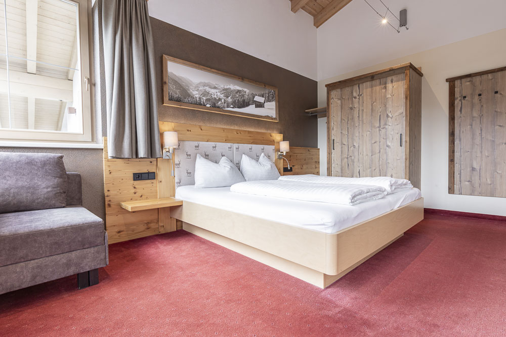 Das Apartment 6 für 4-5 Personen in Ladis verfügt über ein gemütliches Schlafzimmer mit einem großen Bett, weißer Bettwäsche und einem hölzernen Kopfteil. Zwei Nachttischlampen flankieren das Bett und werden durch einen hölzernen Kleiderschrank ergänzt. Der Raum hat einen roten Teppichboden und über dem Fenster hängt ein malerisches Landschaftsfoto mit Vorhängen.