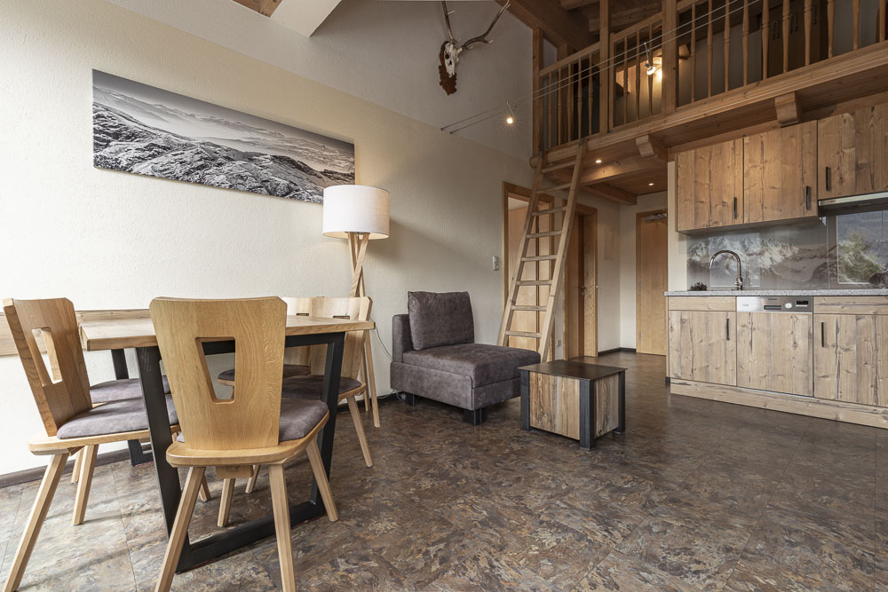 Dieses gemütliche Apartment in Ladis, ideal für 4-5 Personen, verfügt über einen rustikalen Wohnbereich mit Holzmöbeln, darunter ein Esstisch und Stühle, ein grauer Sessel und eine Leiter zum Dachboden. Die Küchenzeile verfügt über einen Herd und eine Spüle, während Landschaftsfotos und montierte Geweihe die Wände zieren.