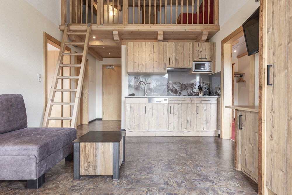Das gemütliche, rustikale Studio-Apartment 6 für 4-5 Personen in Ladis verfügt über einen Holzboden und eine Leiter. Die Küchenzeile ist mit Holzschränken und modernen Geräten ausgestattet. Ein graues Sofa und ein Couchtisch aus Holz stehen auf gemustertem Boden und verstärken das warme, natürliche Ambiente.