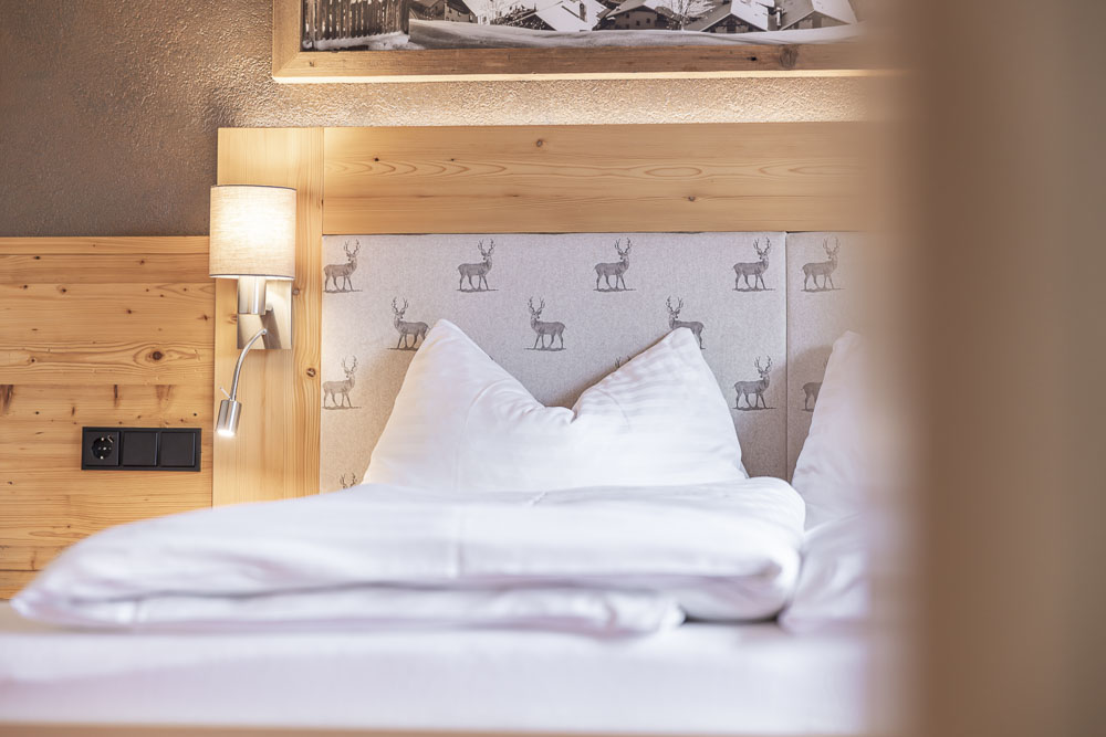 Gemütliches Schlafzimmer im Apartment 6 für 4-5 Personen in Ladis mit einem Kopfteil und Wänden aus Holz. Das Bett ist hübsch mit weißer Bettwäsche bezogen und hat eine Stoffplatte mit kleinen Hirschmustern. Eine Wandlampe mit beigem Lampenschirm erhellt den Raum warm.