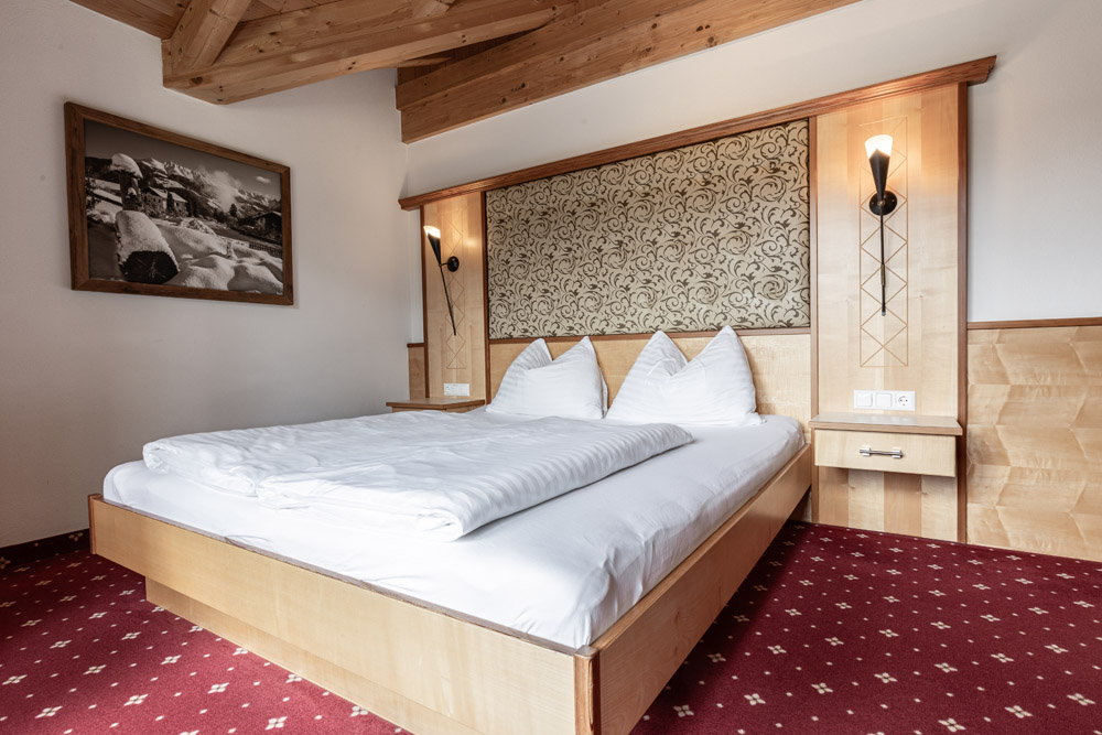 Das gemütliche Apartment Morgensonne in Ladis verfügt über ein Zimmer im Hotelstil mit einem großen Doppelbett mit weißer Bettwäsche und dreieckigen Kissen. Holzvertäfelungen und ein dekoratives Kopfteil schmücken die Wand, während darüber ein Foto hängt. Zwei Nachttische flankieren das Bett. Perfekt für 2-4 Personen.